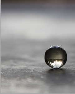 Description: a close-up photo of a colorless translucent sphere resting on cement; in the glass marble is an upside-down reflection of a tree and other indistinct shapes.