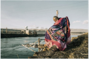 an image of Lana Jack from behind, arms outstretched, facing an industrial site on the river. A photo of Lana Jack from behind, her arms spread out, a butterfly clip in her hair and wearing a black mult-color print, facing an industrial site on the river