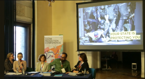 Photo of the panel during Viktoria Kalyniuk's presentation, the slide projected behind is an image of a protester being brutally taken down by heavily armored riot police with the caption "Your State is Protecting You".