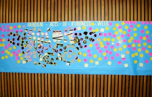 A board that reads "Random Acts Of Kindness Week" with post-it notes on it, with the Self Help Radio logo superimposed in the notes